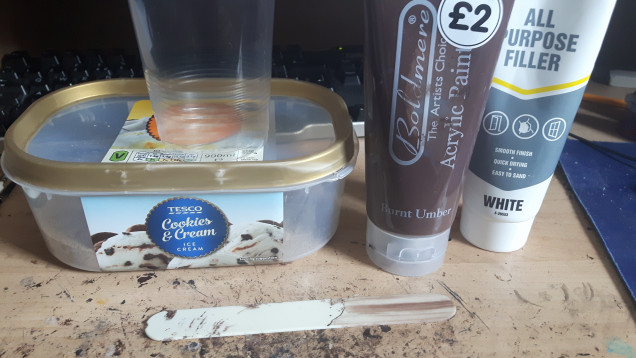 Time to make some texture paint.  Here we have a disposable pint glass to mix it in.  A tongue depresser for stirring.   An ice cream tub full of sand.  Some cheap filler in a squeezy tube and some cheap dark brown paint. 