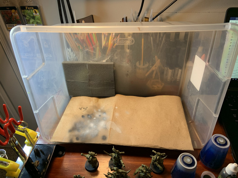 This is my homemade spray booth. It's a plastic tub that I've cut a hole in the bottom and secured a portable battery powered fan to. I then re-used some packing foam from some model kits and glued them over the hole as a filter. This cuts down on overspray quite a bit. Also, the clear container allows light from my desk lamps in. One more bonus is that I pack away my compressor and airbrush kit on top of the upside-down lid, pop the tub/booth over it, and store everything together (again upside-down).